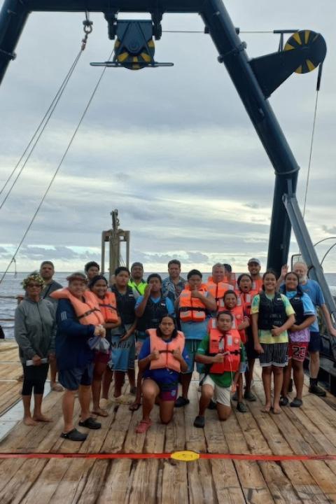 Residents of Mauke, Cook Islands, stand together on the deck of a deep-sea mining boat under a pulley system.